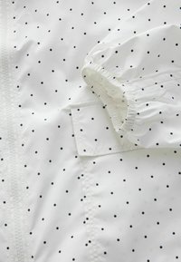 Tissu blanc à petits pois noirs, avec un poignet élastique froncé et une poche rectangulaire à rabat.