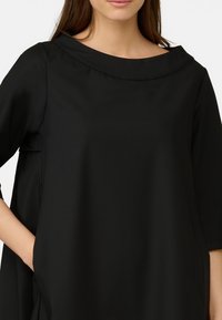 Svart topp med vid, rund halsringning och trekvart ärmar. Tillverkad av ett slätt tyg. Har sidofickor. Enkel, löst sittande design.