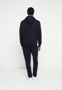 Granatowa bluza z kapturem i dopasowane spodnie dresowe wykonane z miękkiego materiału. Posiada kaptur, prążkowane mankiety oraz białe tenisówki. Prosta, gładka faktura.