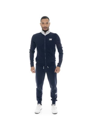 Homme debout portant un survêtement bleu marine avec des logos blancs et des baskets blanches sur un fond blanc uni.