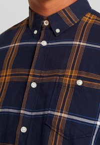 Chemise à carreaux marine avec rayures orange et blanches, en coton doux, col pointu et poche poitrine avec fermeture à bouton.