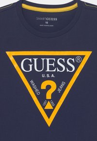 Mørkeblå bomulls t-skjorte med et stort hvitt og gult trekantet grafikk som har teksten "GUESS U.S.A." og "WASHED JEANS", med et sentrert spørsmålstegn.