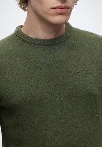 Jeune personne portant un pull en maille vert moucheté avec col côtelé et petit logo brodé sur la poitrine.
