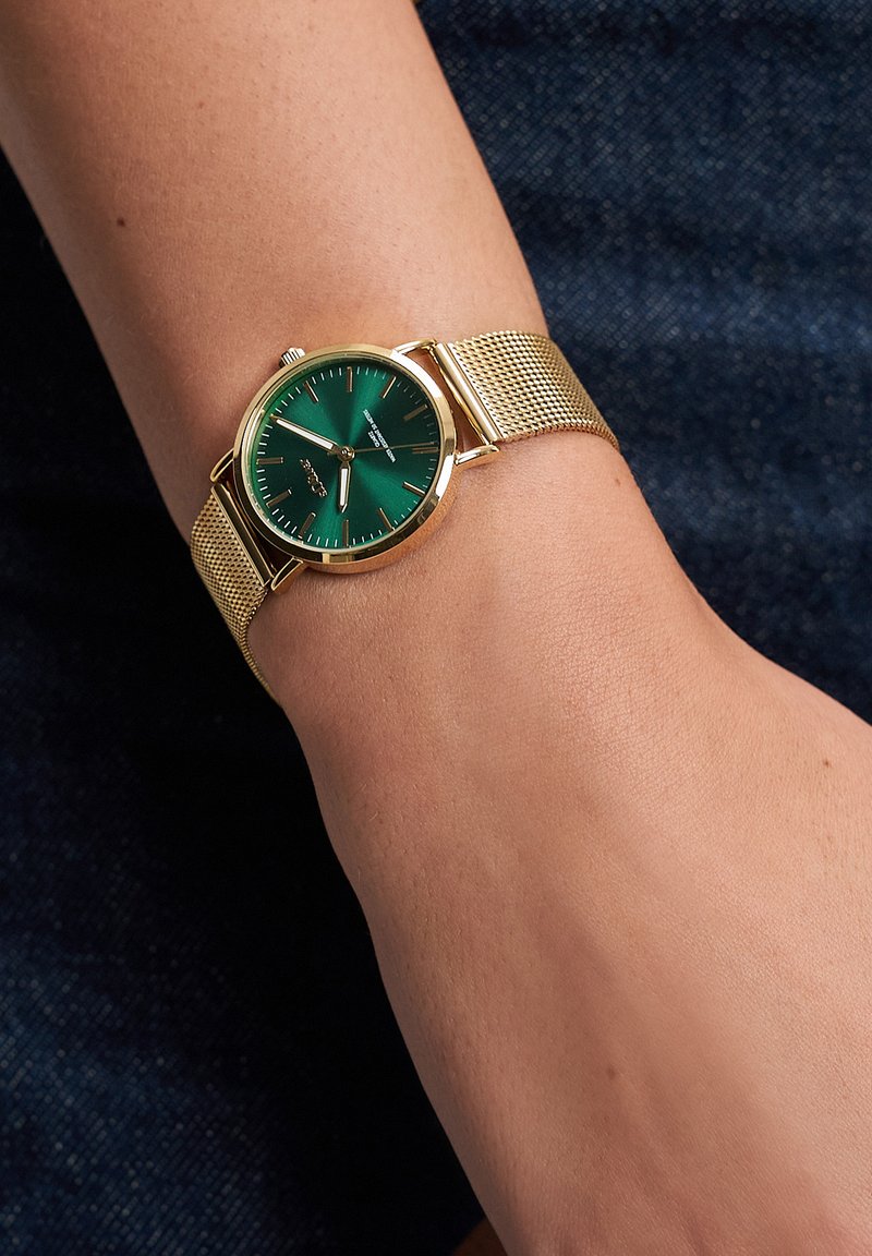 Guldnetrem ur med grøn rund urskive, sølv timeindikatorer og et minimalistisk design. Båret på et håndled mod en denim baggrund.