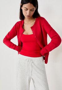 Cardigan rojo de botones sobre una camiseta sin mangas a juego, combinado con pantalones de chándal gris claro. Textura suave, puños y cinturilla acanalados.