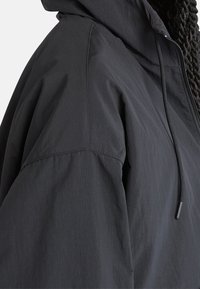 Giacca leggera nera con cappuccio, chiusura frontale con zip, coulisse e tessuto strutturato con un motivo a griglia sottile.