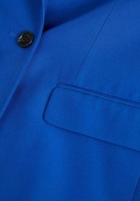 Blå blazer med en slät textur, med en enda svart knapp och en bröstficka med en stilren klaff.