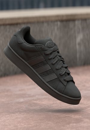 Schwarzer Adidas Campus Sneaker mit Schnürsenkeln, drei Streifen und Logo, schwebend über einer Betonoberfläche und einem Hintergrund mit Steinbank.
