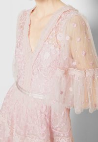 Robe en dentelle rose avec des manches transparentes brodées, des motifs floraux, un décolleté en V et une ceinture en satin. Tissu léger et texturé.