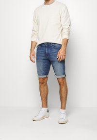 Muž stojí v neformálním oblečení: krémová košile s dlouhým rukávem, modré džíny šortky a bílé tenisky, na pozadí bílé plochy.