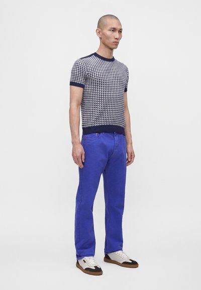 Homme debout portant un haut à manches courtes à motifs géométriques bleu marine et blanc, un pantalon bleu vif et des baskets blanches avec des détails noirs.