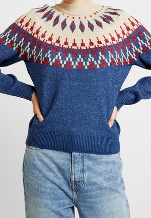 Personne portant un pull en maille bleu avec un empiècement crème orné de motifs géométriques rouges, bleus et blancs, associé à un jean bleu clair.