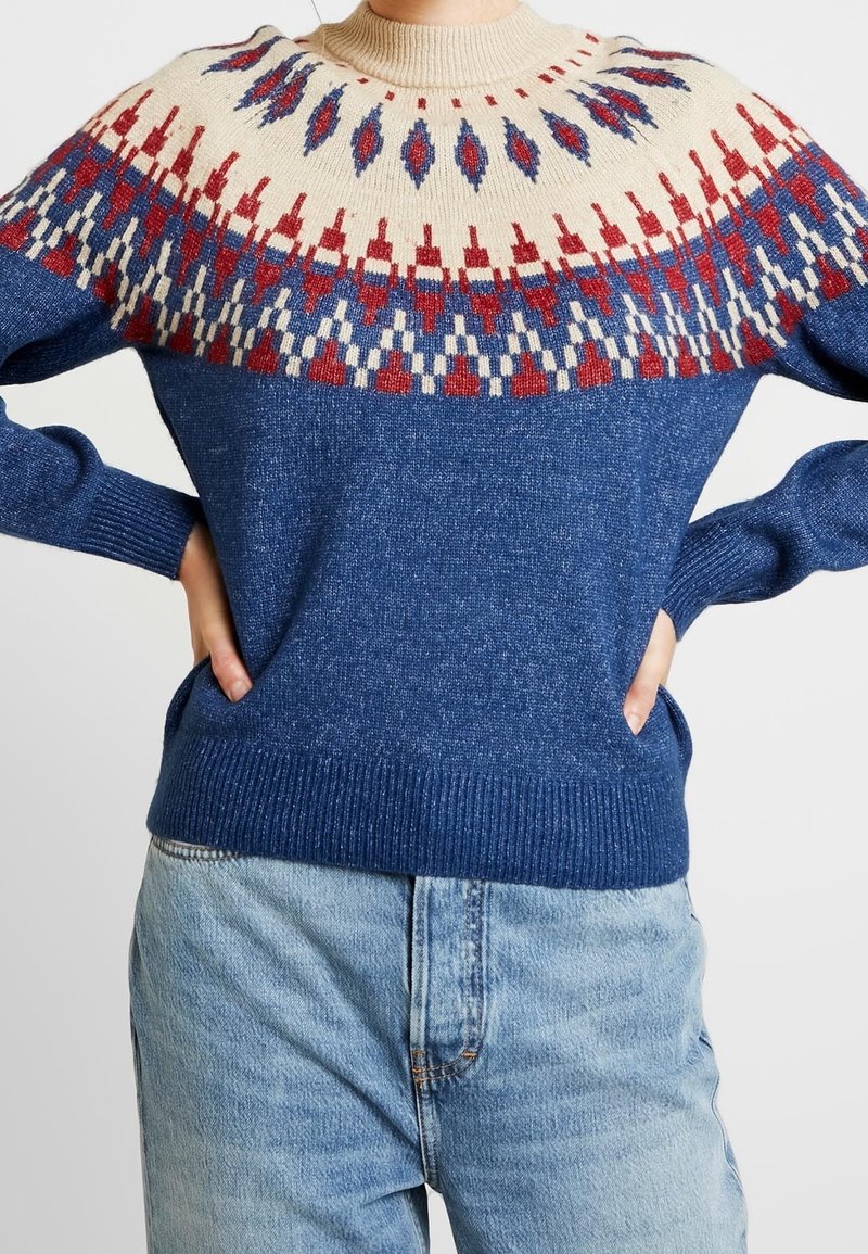 Personne portant un pull en maille bleu avec un empiècement crème orné de motifs géométriques rouges, bleus et blancs, associé à un jean bleu clair.