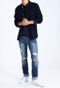 Man i mörkblå jacka över marinblå skjorta, slitna blå jeans uppvikta vid fållarna, och vita sneakers framför vit bakgrund.