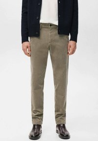 Man draagt beige corduroy broek, zwarte laarzen, wit overhemd en zwarte button-up vest tegen een effen witte achtergrond.