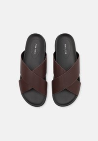 Sandales à brides croisées brun avec une texture lisse et une semelle intérieure noire. Design simple, arborant le nom de la marque sur la semelle.