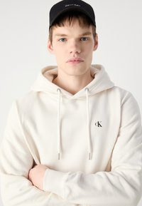 Sudadera blanca con cordones, que presenta un pequeño logo negro "ck". El modelo lleva una gorra negra. Textura suave, diseño casual, mangas largas.