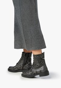 Bottines noires texturées avec un motif reptilien, à lacets et fermeture éclair, avec une semelle en caoutchouc épaisse, associées à un pantalon large gris.