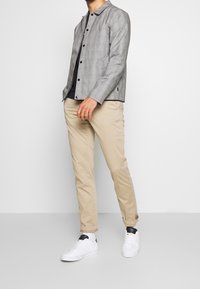 Giacca leggera a quadri grigi con colletto, bottoni neri, abbinata a pantaloni chinos slim-fit beige e sneaker bianche.