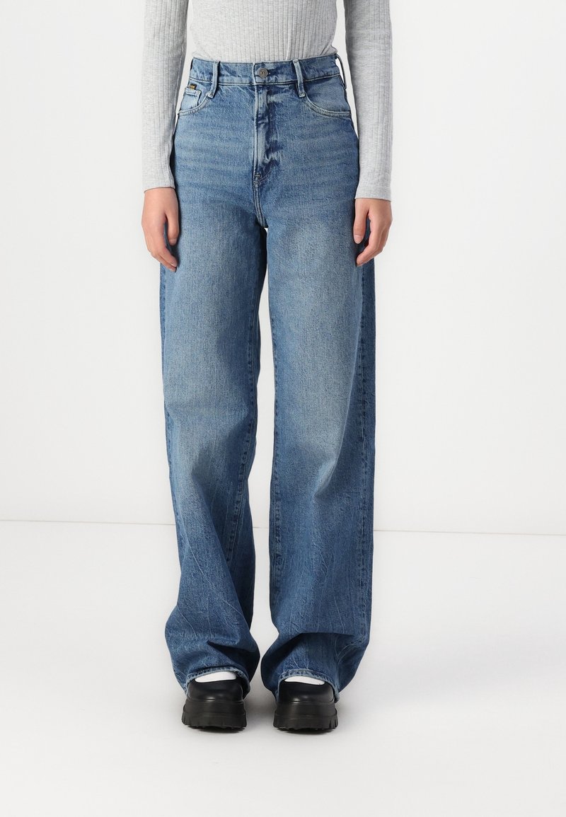 Jeans bleus à jambe large en denim, avec une taille haute, un délavage et des coutures contrastées. Associés à des chaussures de plateforme noires.