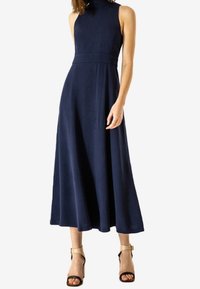Námořnická modrá, bez rukávů maxi šaty s vysokým výstřihem a přiléhavým pasem. Vyrobené z hladké látky, s volánkovou sukní. Černé sandály na vysokém podpatku.
