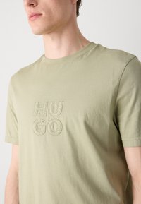 Olive green cotton t-shirt with short sleeves, featuring embroidered "HUGO" logo in darker stitching on the front. Smooth texture.