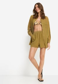 Πουκάμισο σε ελιά πράσινο με κουμπιά, φορεμένο πάνω από μπικίνι με λεοπάρ σχέδιο και σορτς με το ίδιο μοτίβο, που διαθέτουν ελαστική μέση και πλαϊνές τσέπες. Μαύρα σανδάλια με επίπεδη σόλα.