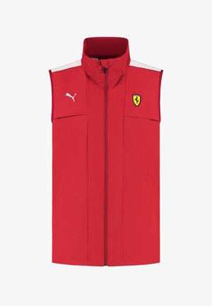 Czerwona bezrękawnik wykonany z lekkiego materiału, z wysokim kołnierzem, zamkiem błyskawicznym z przodu, białymi akcentami i logo Ferrari po lewej stronie klatki piersiowej.