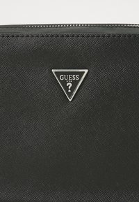 Zwarte portemonnee met een gestructureerd oppervlak, voorzien van een zilveren driehoekig GUESS-logo en een ritsluiting. Compact ontwerp voor gemakkelijke draagbaarheid.
