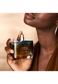 Person som holder en Bronze Goddess Estée Lauder-parfymeflaske nær nakken, med lett smilende lepper og iført gullfargede hoop-øreringer.