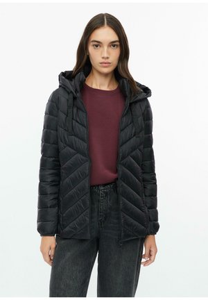 Frau trägt eine schwarze gesteppte Kapuzenjacke über einem bordeauxfarbenen Shirt und dunkelblauen Jeans, steht vor einem schlichten weißen Hintergrund.