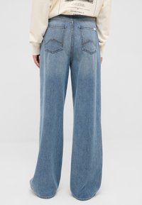 Persona che indossa jeans a gamba larga azzurri e una felpa color crema, vista da dietro su uno sfondo semplice.