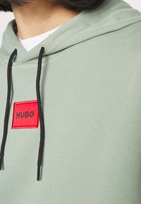Felpa verde chiaro con un ampio patch rettangolare rosso etichettato "HUGO". Presenta corde nere e una texture a coste sul tessuto.