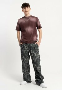 Gładka, ciemna bordowa koszulka połączona z szarymi spodniami w luźnym kroju w kamuflażowy wzór oraz białymi sneakersami. Model stoi, trzymając ręce w kieszeniach.