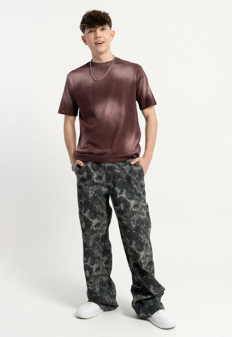 Gładka, ciemna bordowa koszulka połączona z szarymi spodniami w luźnym kroju w kamuflażowy wzór oraz białymi sneakersami. Model stoi, trzymając ręce w kieszeniach.