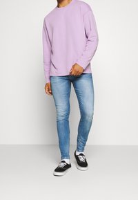 Camisa de manga larga color púrpura claro, vaqueros ajustados azules y zapatillas negras. El conjunto presenta un diseño sencillo y materiales casuales.