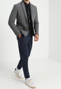 Blazer gris avec revers crantés, fermeture à un bouton, associé à une chemise noire et un pantalon bleu marine. Baskets blanches avec des accents de design minimal.