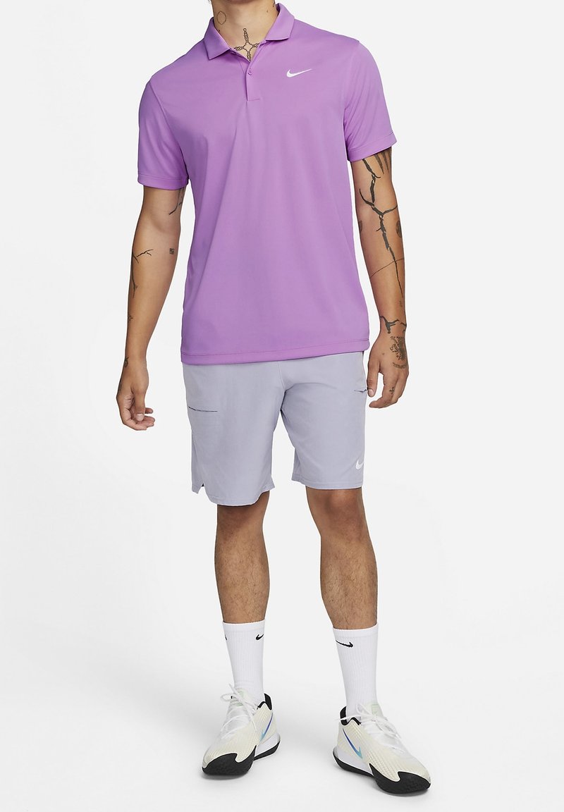 Mężczyzna w lawendowej koszulce polo Nike, jasnoszarych szortach Nike, białych skarpetkach Nike oraz białych butach sportowych, stoi na białym tle.