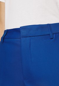 Närbild av blå skräddarsydda byxors midja bärs med en vit insydd skjorta, som visar bältesöglor och detaljer i framsömnaden.