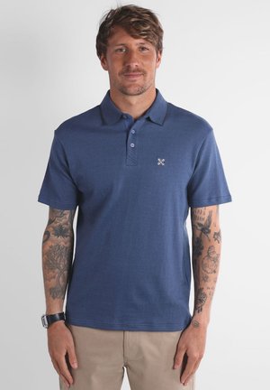 Homme aux cheveux châtain clair portant un polo bleu à manches courtes et un pantalon beige, affichant des tatouages sur les deux avant-bras et une montre au poignet gauche.