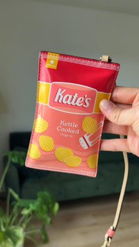 Röd snacksäck med gula grafik av kettle-cooked potatischips, "Kate's" logotyp och en illustration av en saltkar. Slät, glänsande yta, med ett bärrem.