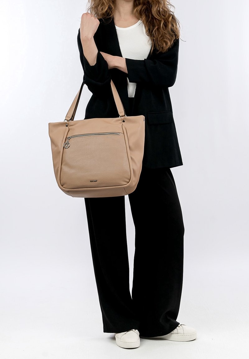 Femme portant une tenue noire et des chaussures blanches tenant un grand sac fourre-tout beige avec une poche zippée à l'avant sur un fond uni.