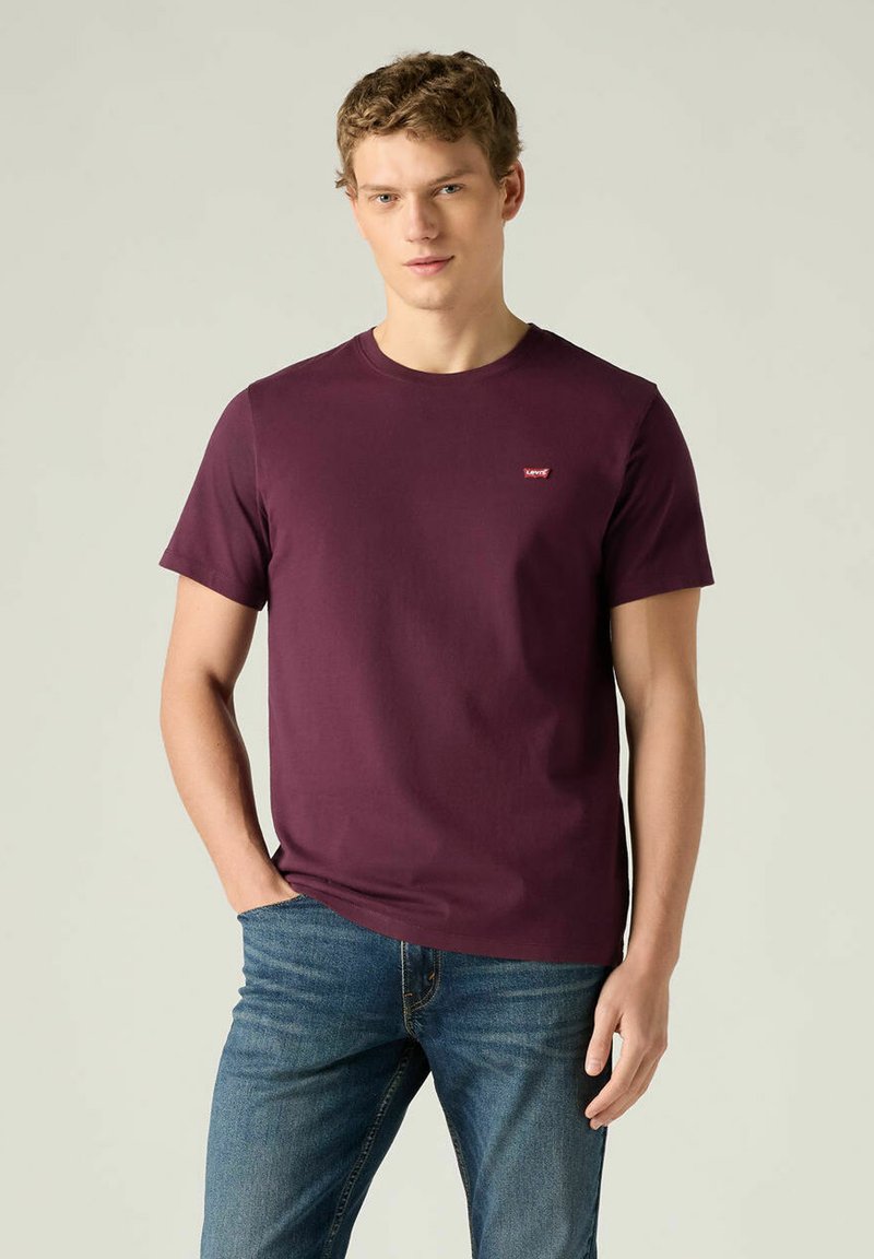 Junger Mann trägt ein schlichtes burgunderrotes T-Shirt mit kleinem Logo auf der Brust, kombiniert mit blauen Jeans, steht mit einer Hand in der Tasche.