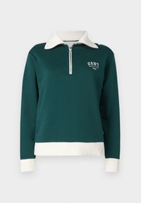 Pulóver verde con cuello y dobladillo de canalé blanco, con cremallera en la parte frontal y logotipo en el pecho. Textura de tela suave.