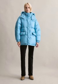 Ljusblå vadderad jacka med hög krage, designad med stora quiltade sektioner, två framfickor och en avtagbar huva.