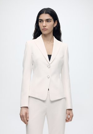 Vrouw met lang donker haar draagt een nauwsluitend wit colbert met twee knopen aan de voorkant en bijpassende witte broek tegen een effen achtergrond.