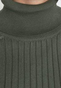 Dunkelgrüner, gerippter Strickpullover mit Rollkragen, der einen hohen Kragen und strukturierte, vertikale Linien im gesamten Stoff aufweist.