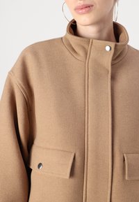 Cappotto color cammello con colletto alto, realizzato in tessuto testurizzato. Presenta due tasche frontali con chiusure a scatto e un design pulito e strutturato.