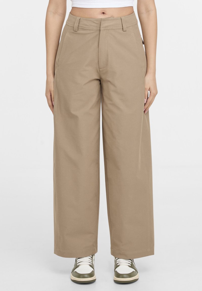 Pantalon beige taille haute à jambes larges avec poches avant, porté avec des baskets blanches et vertes et un haut court blanc.