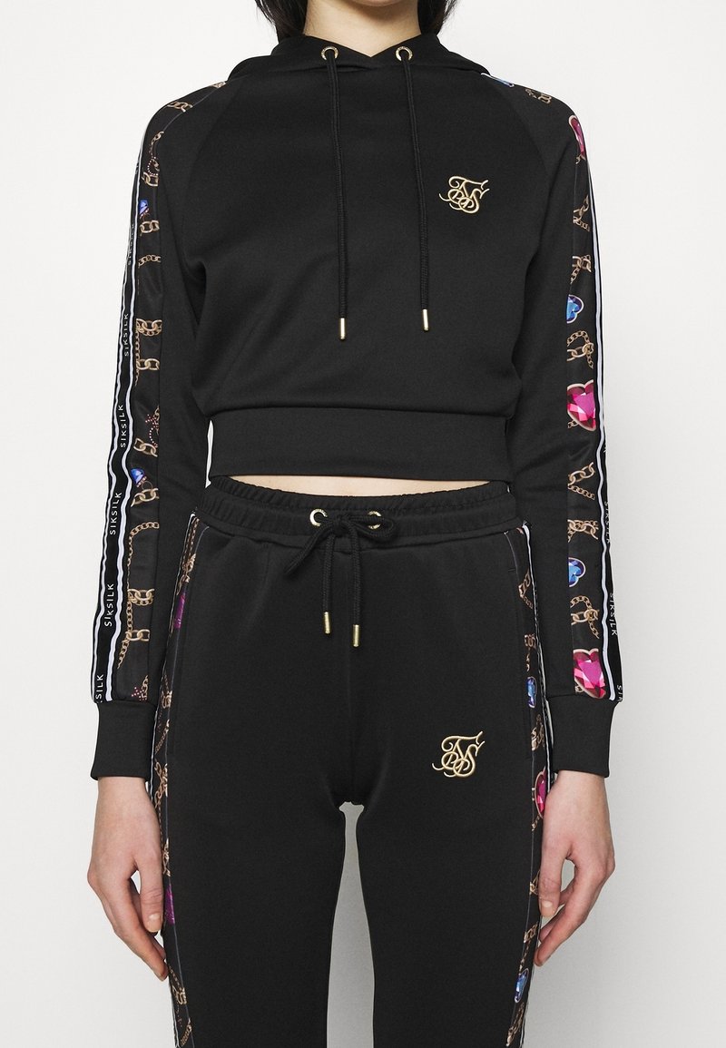 Sweat à capuche court noir et joggings assortis avec logo doré et motif coloré de chaînes et de pierres précieuses le long des manches et des côtés.